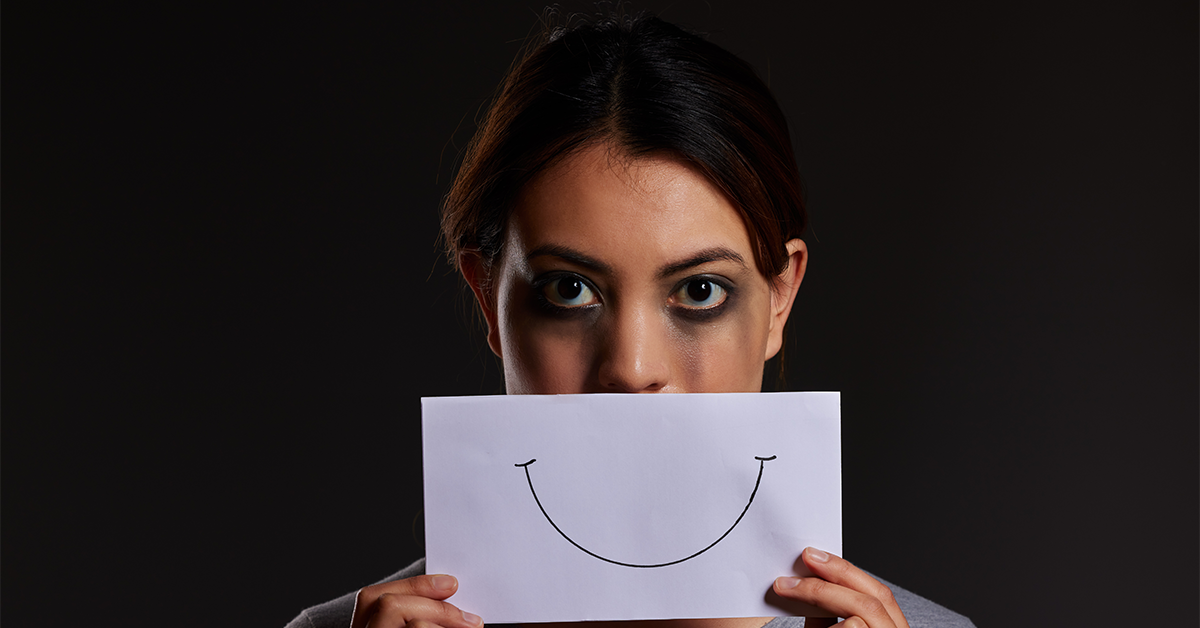Woman with smeared eye makeup hiding her mouth behind a paper with a drawn smile.