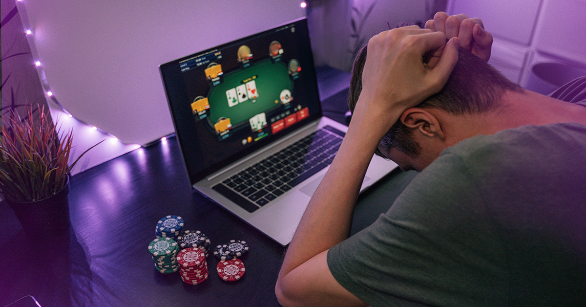 Person with head in hands watching online poker on a laptop; poker chips stacked on the desk nearby.